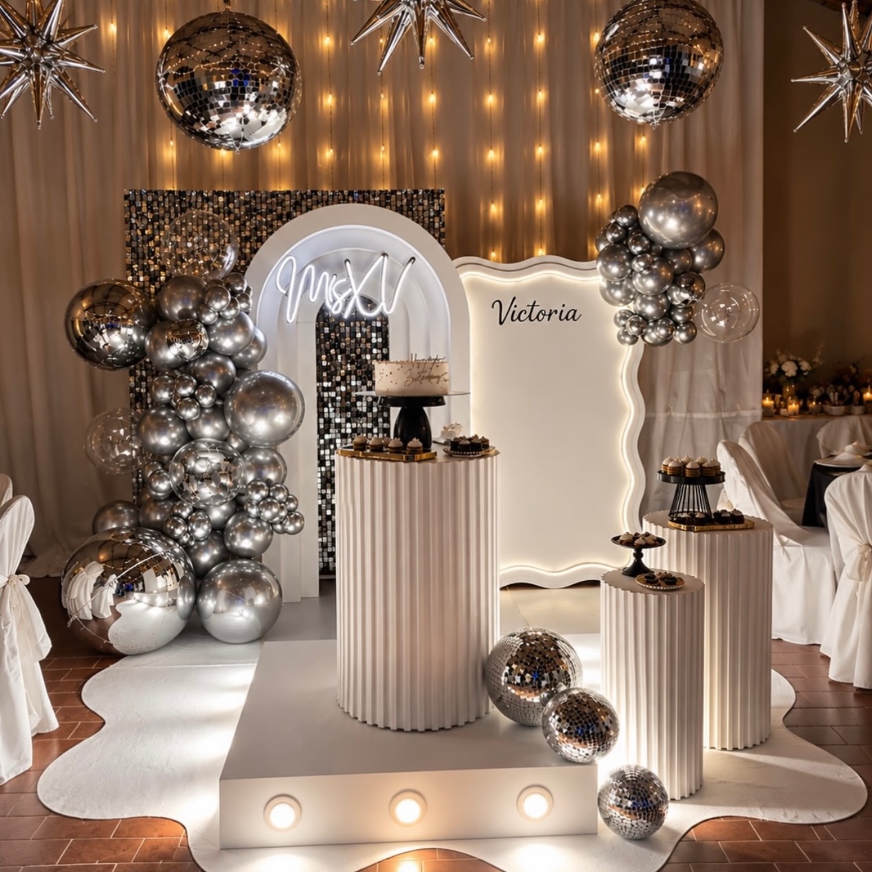 Montaje de quince años en blanco y plata con arco iluminado, globos cromados y pedestales
